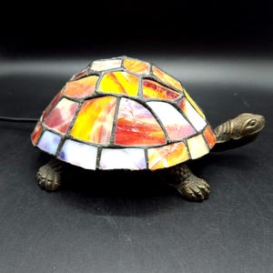 May include: A stained glass turtle lamp with a shell in red, orange, yellow, and white. The turtle's body and legs are bronze-coloured. The lamp is designed to sit on a surface and has a black power cord.