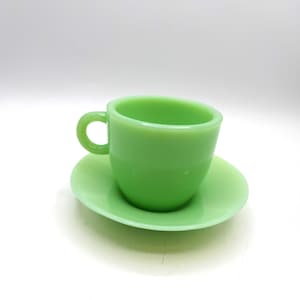 May include: A light green teacup and saucer set. The cup has a rounded handle and a cylindrical shape. The saucer is round and flat. Both pieces are made of a solid, opaque material.
