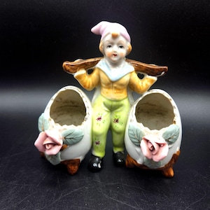 May include: A vintage ceramic figurine featuring a young figure in a yellow jacket, green pants, and a pink hat, carrying a brown beam. Two egg-shaped planters with pink roses and green leaves flank the figure. The figurine is on a black surface.