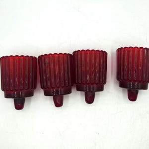 May include: Four red glass candle holders with a ribbed design. Each holder has a scalloped top and a small, rounded base. The candle holders are arranged in a row against a white background.