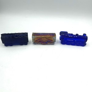 May include: Three miniature glass train carriages. One is a dark blue passenger carriage, another is a brown and gold freight carriage, and the third is a cobalt blue locomotive. The carriages are displayed on a white surface.