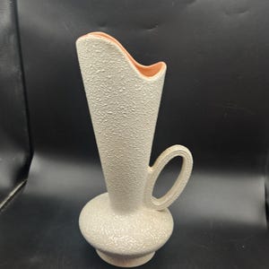 May include: A tall, off-white ceramic vase with a textured, pebbled exterior finish. The vase features a wide, rounded base, a tapered neck, and a single side handle. The interior rim and opening are finished in a smooth, solid peach-colored glaze.