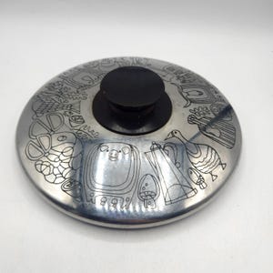 May include: A stainless steel pot lid with a black knob. The lid is decorated with etched line drawings of various objects, including a pig, a teapot, a duck, and musical instruments. The lid is round.