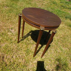 May include: A small side table with a circular dark brown wood grain top and four tapered brown legs. The legs are connected to the tabletop by metallic gold-colored hardware accents. The table features a simple construction with a flat surface.