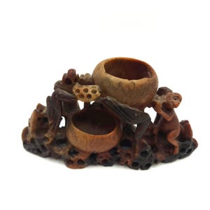 May include: A carved wooden sculpture featuring three open bowls and a monkey figure. The sculpture is a warm brown color with intricate details, including carved floral and leaf patterns. The base is dark brown and textured.