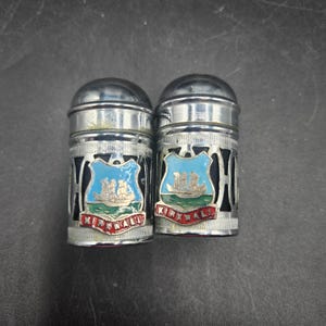 May include: A pair of vintage salt and pepper shakers with a silver finish. Each shaker features a blue and white crest with a ship design and the word "KIRKWALL" in red. The shakers have a cylindrical shape with a domed top.