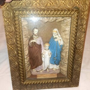 May include: A framed religious relief sculpture of the Holy Family. The figures are in shades of brown, blue, and white, set against a neutral backdrop. The ornate gold frame adds a decorative touch.