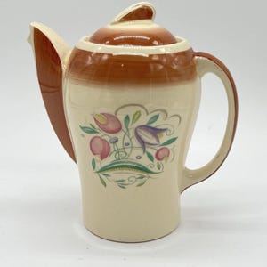 May include: Cream-colored ceramic teapot with a brown lid and handle. The teapot features a floral design with pink, purple, and green accents. The spout is a contrasting brown color.