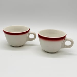 May include: Two white ceramic coffee cups with a red rim and handle. The cups are placed side by side on a white surface. The cups are empty and ready to be filled with coffee or tea.