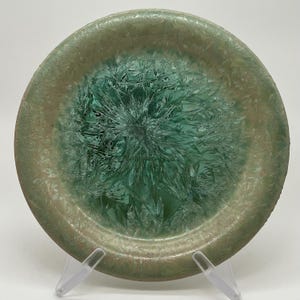 May include: A round, decorative plate with a unique, textured design. The plate features a central burst of teal-green, surrounded by a rim of muted green and brown. The intricate pattern resembles a crystallized or fractal design, adding visual interest.