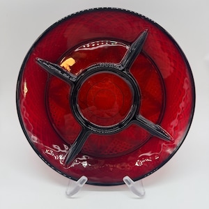 May include: A ruby red glass serving dish with a divided design. The dish has a circular shape with a central compartment and four radiating sections, all outlined in black. The dish is supported by a clear stand.