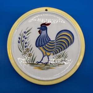 May include: Round ceramic wall plate with a hand-painted rooster design. The rooster is blue and yellow, with a red comb, set against a white background. The plate has a yellow border and a small hole for hanging.