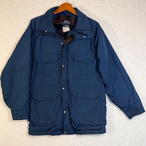 May include: A navy blue jacket with a collared neckline and multiple front pockets. The jacket has a button-up closure and a plaid lining visible at the collar. The jacket is hanging on a wooden hanger.