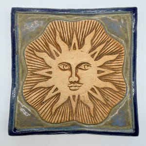 May include: A square ceramic tile featuring a central embossed sun motif with a human face and radiating rays. The sun is unglazed tan clay, while the surrounding background is a muted sage green with a dark blue border framing the entire piece.