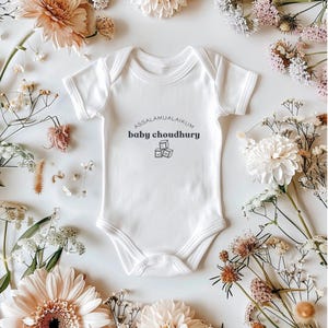 May include: White baby onesie with the text "ASSALAMUALAIKUM baby choudhury" and a graphic of three stacked blocks. The onesie is surrounded by an assortment of flowers in shades of white, cream, and peach.
