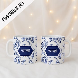 May include: Two white ceramic mugs featuring a blue and white floral pattern. Each mug has a central dark blue rectangular label with rounded corners. The left mug displays the Bengali text 'আম্মা' and the right mug displays 'আব্বা'.