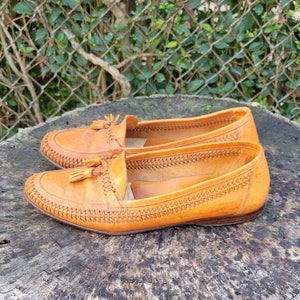May include: A pair of brown leather tassel loafers with a woven pattern on the upper. The loafers are on a wooden surface.