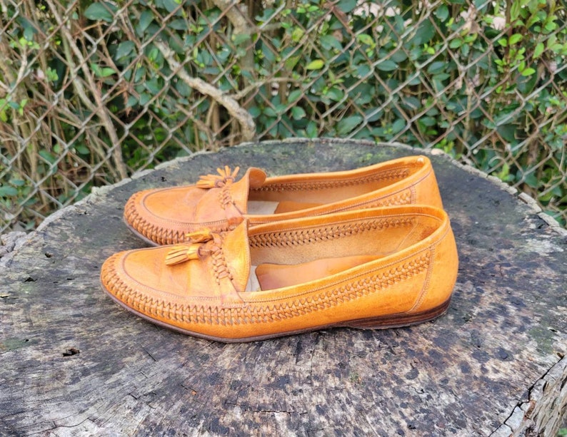 May include: A pair of brown leather tassel loafers with a woven pattern on the upper. The loafers are on a wooden surface.