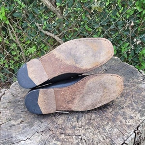 May include: A pair of black leather shoes with worn leather soles. The shoes are placed on a rough wooden surface.