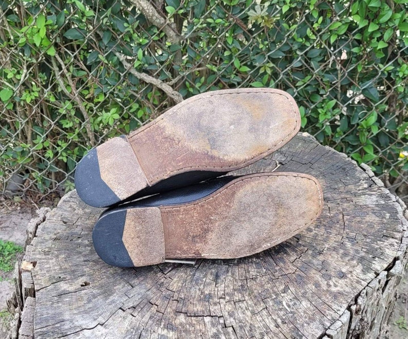 May include: A pair of black leather shoes with worn leather soles. The shoes are placed on a rough wooden surface.