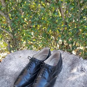 Puede incluir: Un par de zapatos Oxford de cuero negro con detalles de punta de ala. Los zapatos est&aacute;n atados y tienen un acabado pulido.