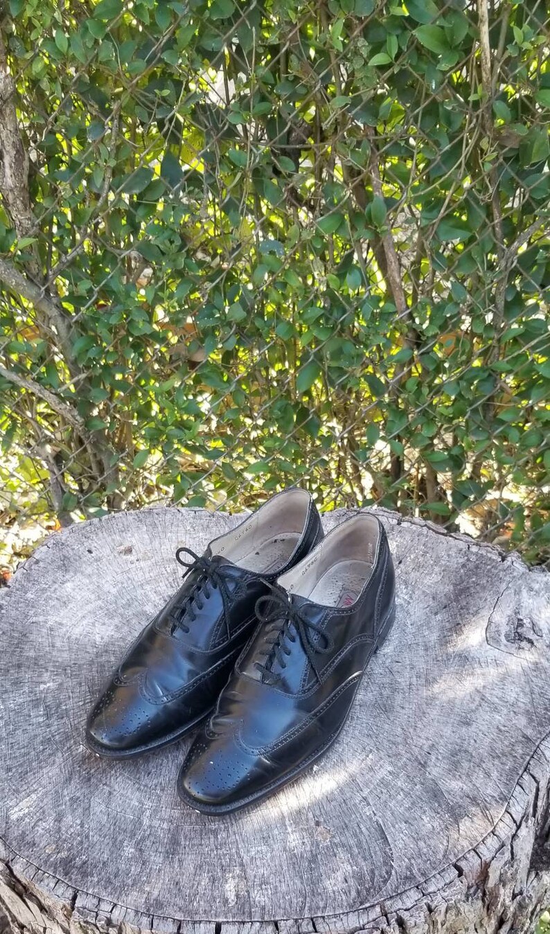 Puede incluir: Un par de zapatos Oxford de cuero negro con detalles de punta de ala. Los zapatos est&aacute;n atados y tienen un acabado pulido.