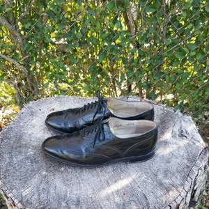 Puede incluir: Un par de zapatos oxford de cuero negro con detalles de punta de ala. Los zapatos tienen cordones y est&aacute;n sobre un toc&oacute;n de &aacute;rbol.