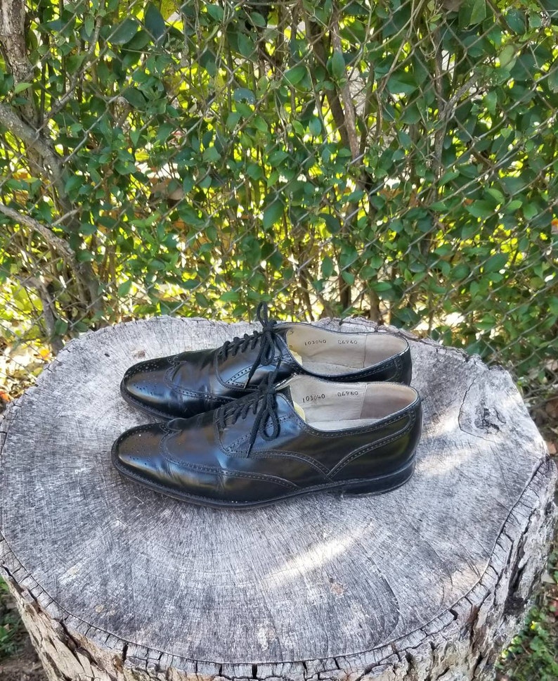 Puede incluir: Un par de zapatos oxford de cuero negro con detalles de punta de ala. Los zapatos tienen cordones y est&aacute;n sobre un toc&oacute;n de &aacute;rbol.