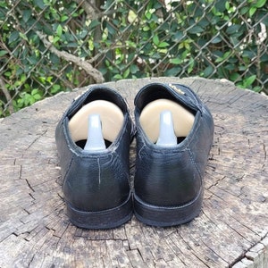 May include: A pair of black leather loafers with gold accents. The loafers have a rounded toe and a low heel. They are sitting on a wooden surface.