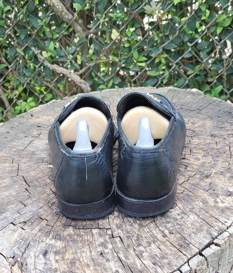 May include: A pair of black leather loafers with gold accents. The loafers have a rounded toe and a low heel. They are sitting on a wooden surface.