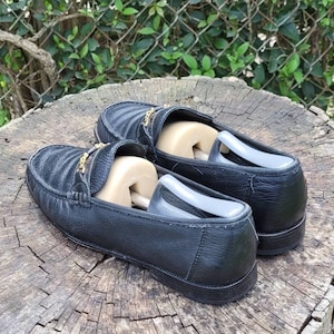 May include: A pair of black leather loafers with gold accents. The loafers have a quilted design and are sitting on a wooden surface.