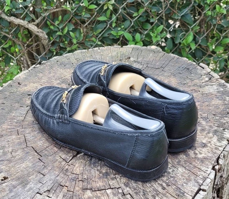 May include: A pair of black leather loafers with gold accents. The loafers have a quilted design and are sitting on a wooden surface.