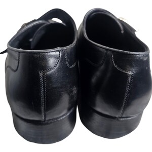 Pode incluir: Um par de sapatos de couro preto com fivelas. Os sapatos t&ecirc;m bico fino e salto baixo.