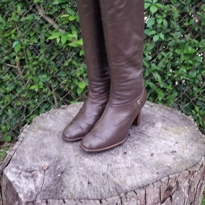 May include: A pair of brown leather knee-high boots with a small heel. The boots are sitting on a tree stump.