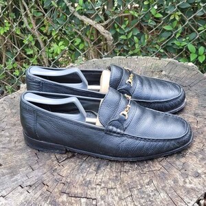 May include: A pair of black leather loafers with gold buckles. The loafers are on a wooden surface.