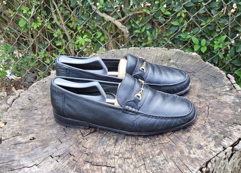 May include: A pair of black leather loafers with gold buckles. The loafers are on a wooden surface.