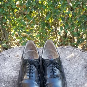 Puede incluir: Un par de zapatos negros de cuero con cordones y detalles de punta de ala. Los zapatos tienen un patr&oacute;n de brogue y est&aacute;n sobre una superficie de madera.