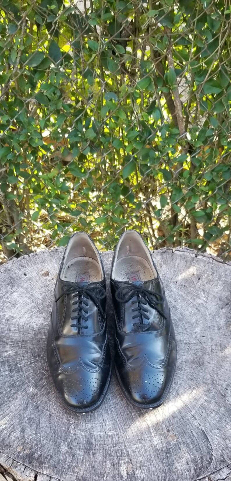 Puede incluir: Un par de zapatos negros de cuero con cordones y detalles de punta de ala. Los zapatos tienen un patr&oacute;n de brogue y est&aacute;n sobre una superficie de madera.