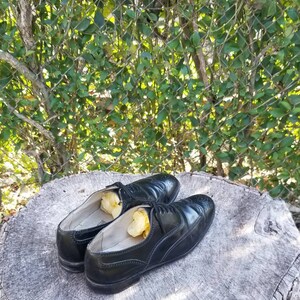 Puede incluir: Dos zapatos de cuero negro con detalles de punta de ala y cordones. Los zapatos est&aacute;n sobre un toc&oacute;n de &aacute;rbol y tienen papel de seda amarillo en su interior.