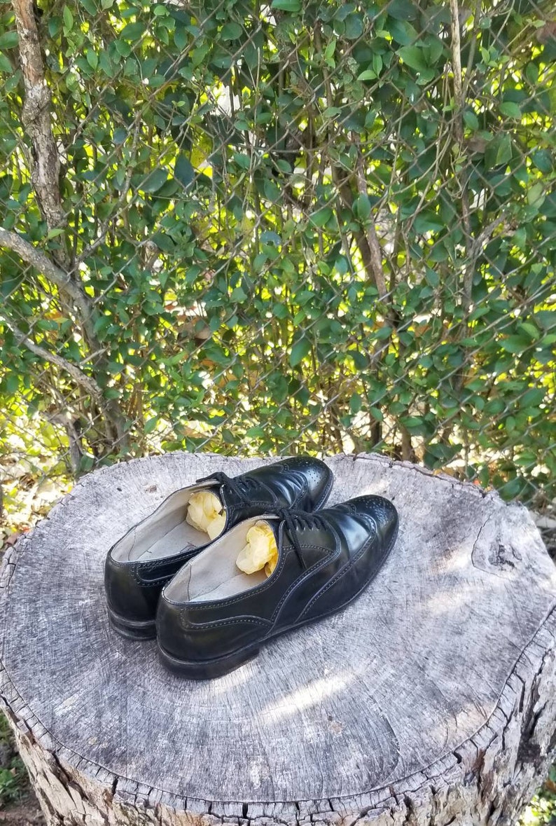 Puede incluir: Dos zapatos de cuero negro con detalles de punta de ala y cordones. Los zapatos est&aacute;n sobre un toc&oacute;n de &aacute;rbol y tienen papel de seda amarillo en su interior.