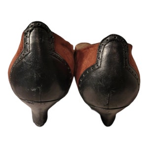 May include: A pair of brown and black suede heeled shoes with a pointed toe. The shoes have a black leather trim and a small heel.