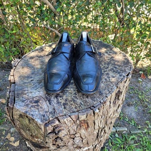 K&ouml;nnte beinhalten: Ein Paar schwarze Lederschuhe mit Schnallenverschluss. Die Schuhe stehen auf einem Baumstumpf.