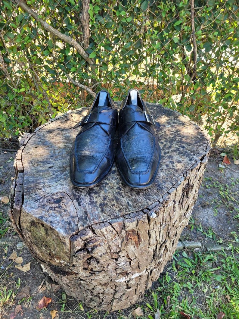 K&ouml;nnte beinhalten: Ein Paar schwarze Lederschuhe mit Schnallenverschluss. Die Schuhe stehen auf einem Baumstumpf.