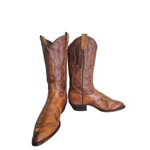 May include: A pair of two-toned brown leather cowboy boots featuring a pointed toe, a stacked heel, and decorative stitching on the shaft. The boots have a lighter tan leather foot and a darker brown leather upper shaft with pull tabs at the top.