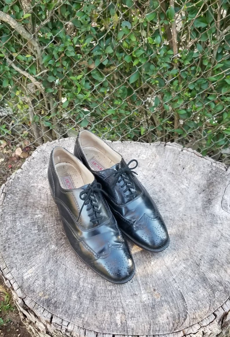 Puede incluir: Un par de zapatos negros de cuero con cordones y detalles de punta de ala. Los zapatos tienen un patr&oacute;n de brogue y est&aacute;n sobre un toc&oacute;n de &aacute;rbol.