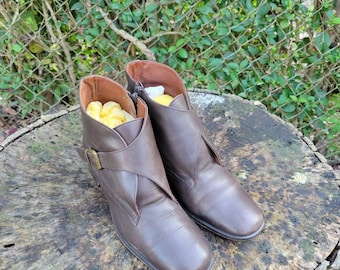 May include: A pair of brown leather ankle boots with a buckle strap on one boot. The boots are on a wooden surface.