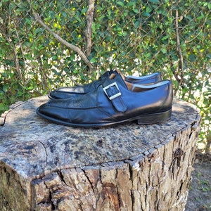 K&ouml;nnte beinhalten: Ein Paar schwarze Lederschuhe mit einem einzigen Schnallenriemen. Die Schuhe stehen auf einem Baumstumpf.