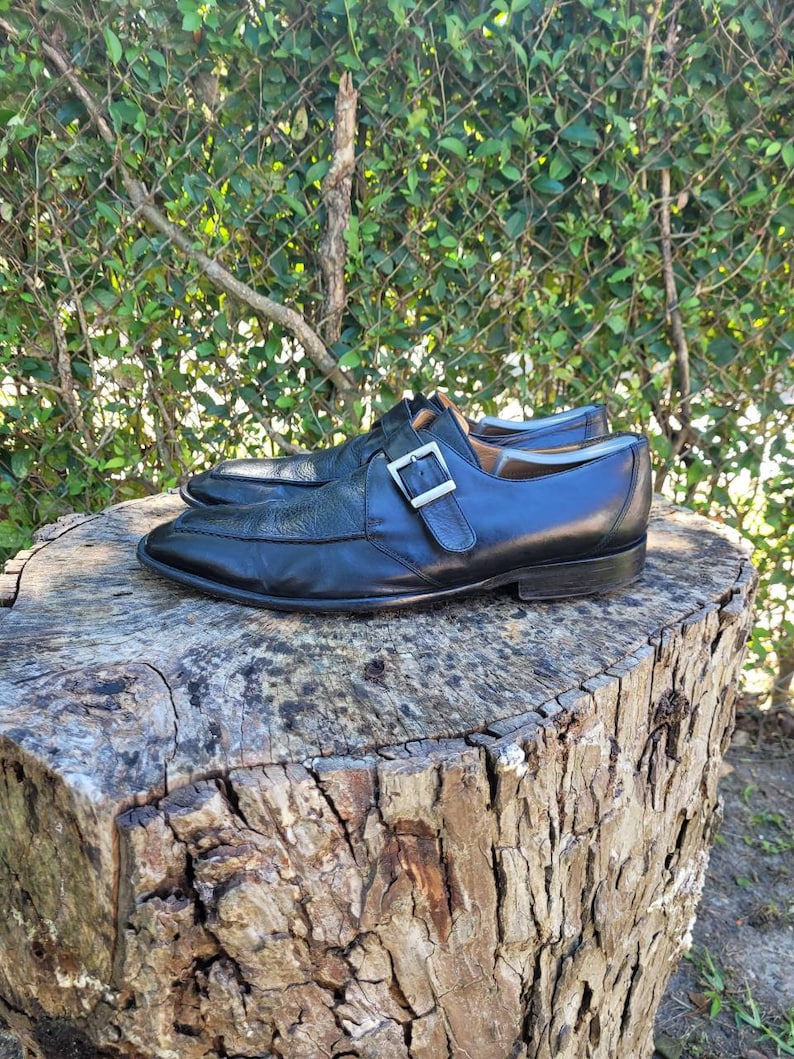 K&ouml;nnte beinhalten: Ein Paar schwarze Lederschuhe mit einem einzigen Schnallenriemen. Die Schuhe stehen auf einem Baumstumpf.