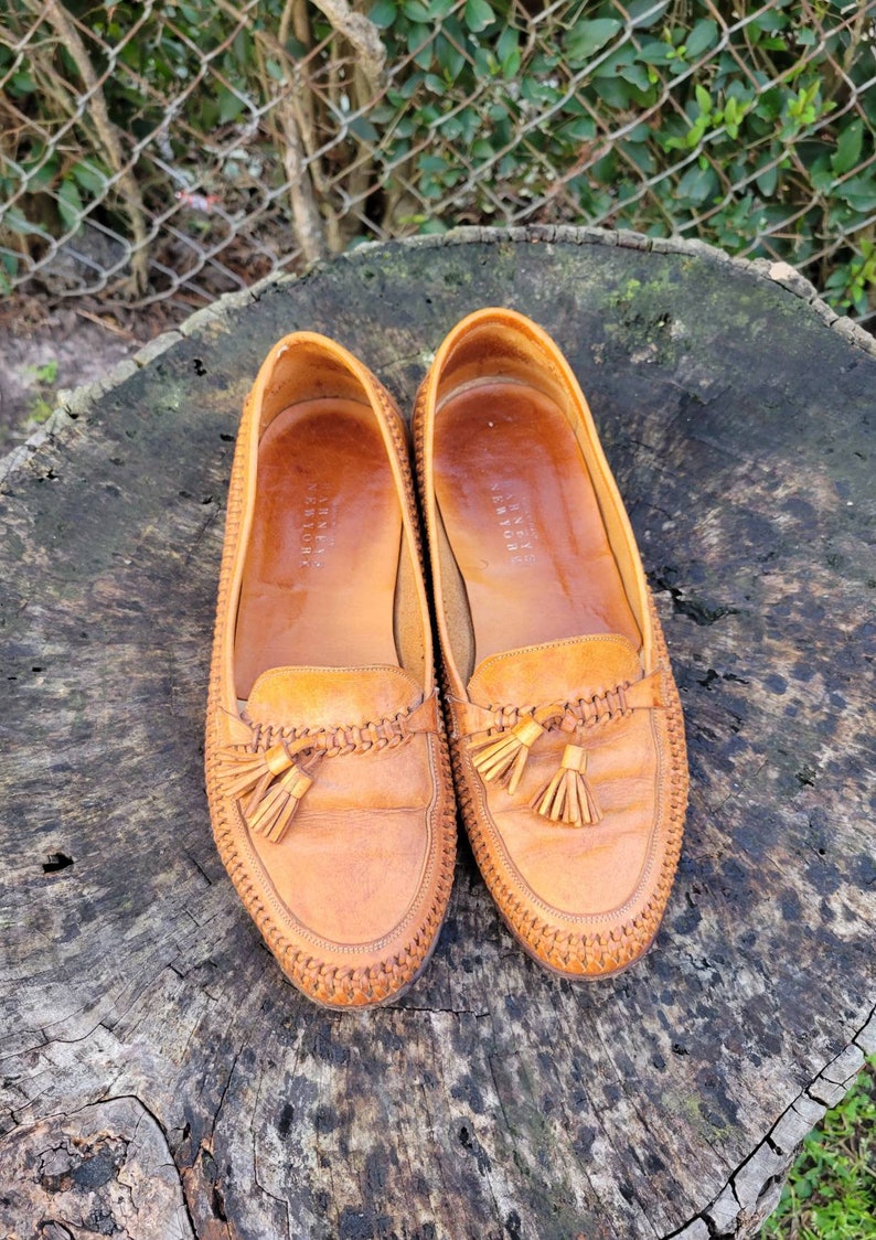 May include: A pair of brown leather tassel loafers with a woven detail around the edge. The loafers are on a wooden surface.