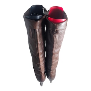May include: A pair of brown leather knee-high boots with a pointed toe. The boots are folded down at the top, revealing a red and black shoe stretcher inside.